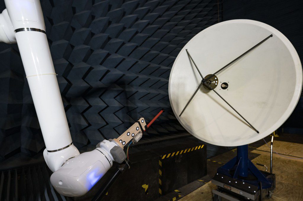 In a testing facility, a robotic arm manipulates a probe to test the electric field of a large, white, circular dish antenna. This antenna is situated within an anechoic chamber, which is lined with dark blue, wedge-shaped foam.