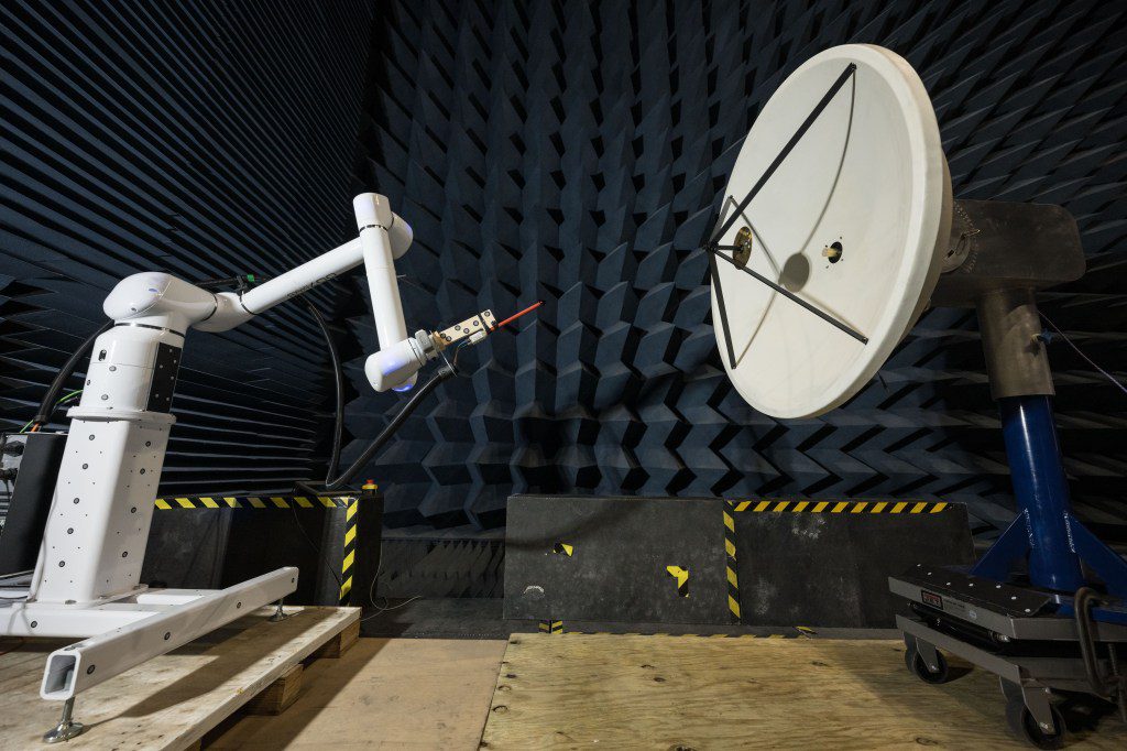 In a testing facility, a robotic arm manipulates a probe to test the electric field of a large, white, circular dish antenna. This antenna is situated within an anechoic chamber, which is lined with dark blue, wedge-shaped foam.