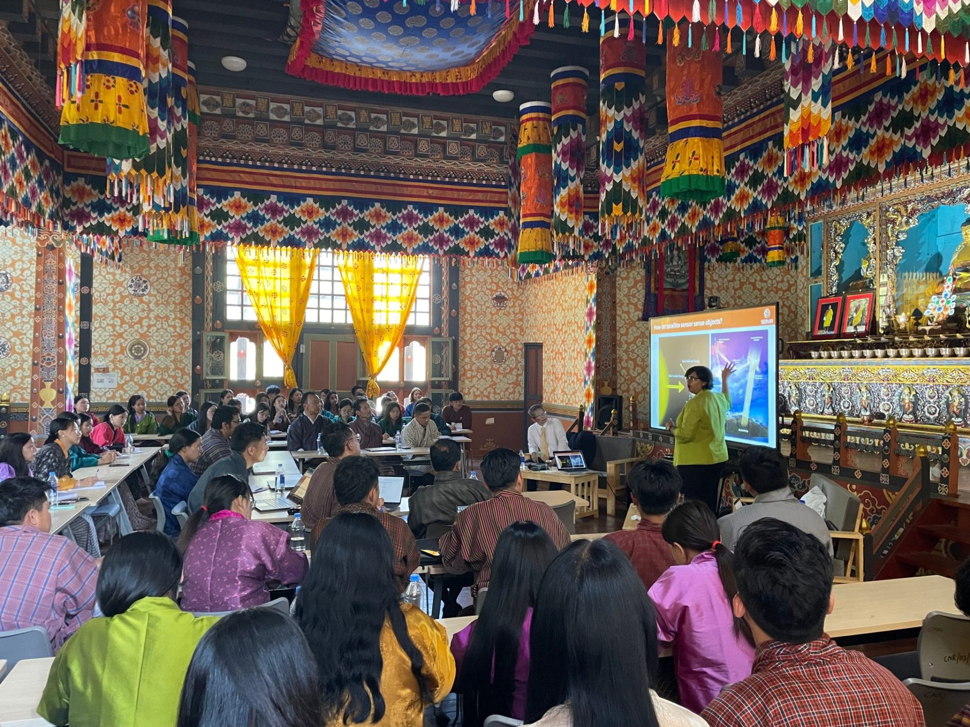 A large group of people sit at tables watching someone present at the Royal University of Bhutan College of Natural Resources.