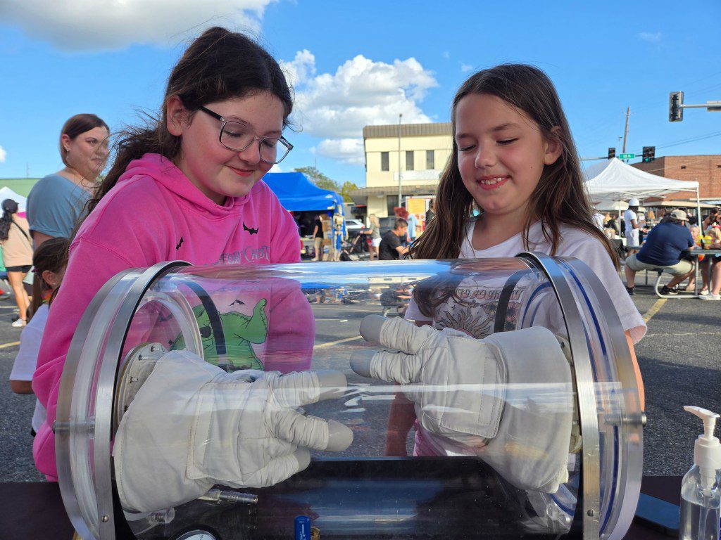 Two members of Artemis Generation at the Picayune Street Fair interact with astronaut gloves on display