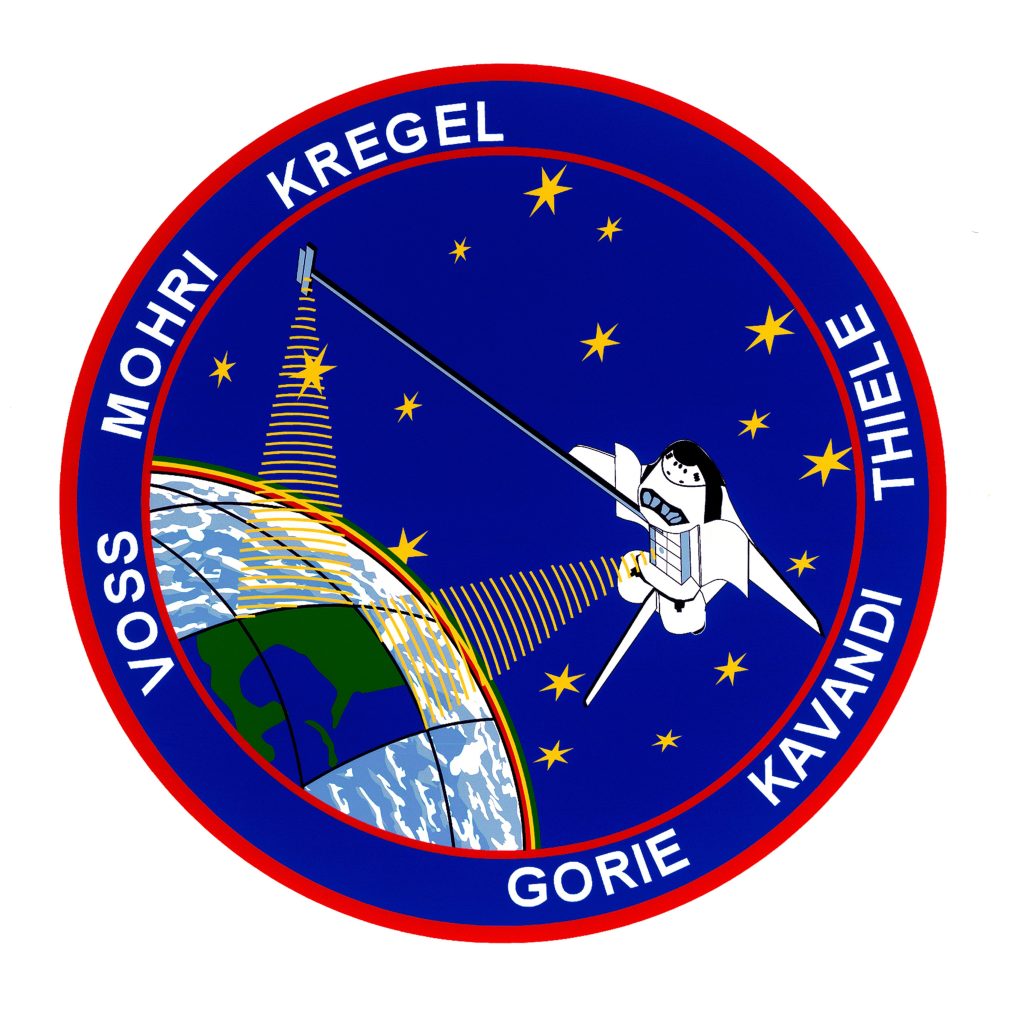 Circular crew patch with a blue background, including a partial Earth and a space shuttle orbiting it, with the names of the six astronauts written along the outer border.